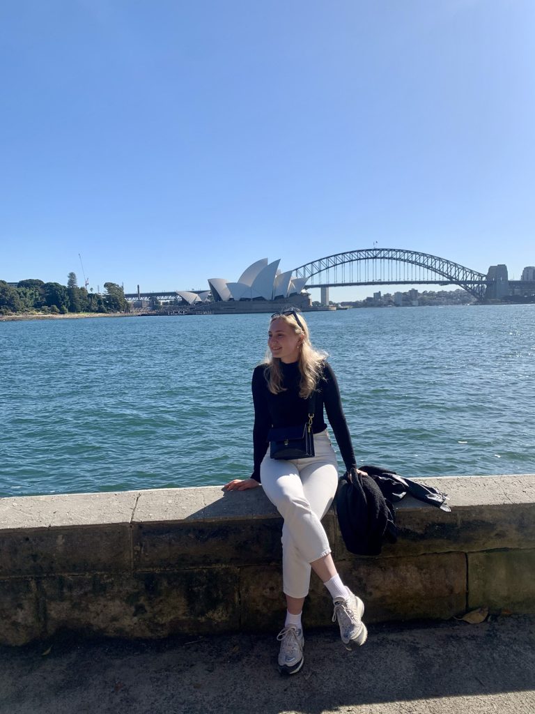 Silva Heinonen istuu merenrannalla muurin päällä ja takana näkyy Sydneyn kuuluisa silta