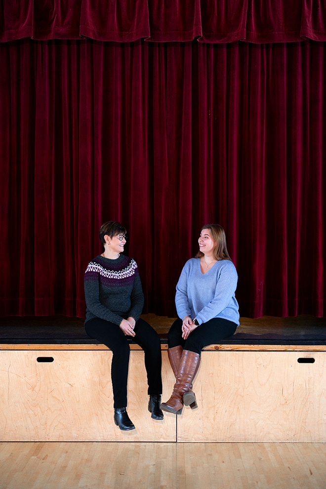 Kaksi naista istuu esiintymislavan reunalla verhojen edessä